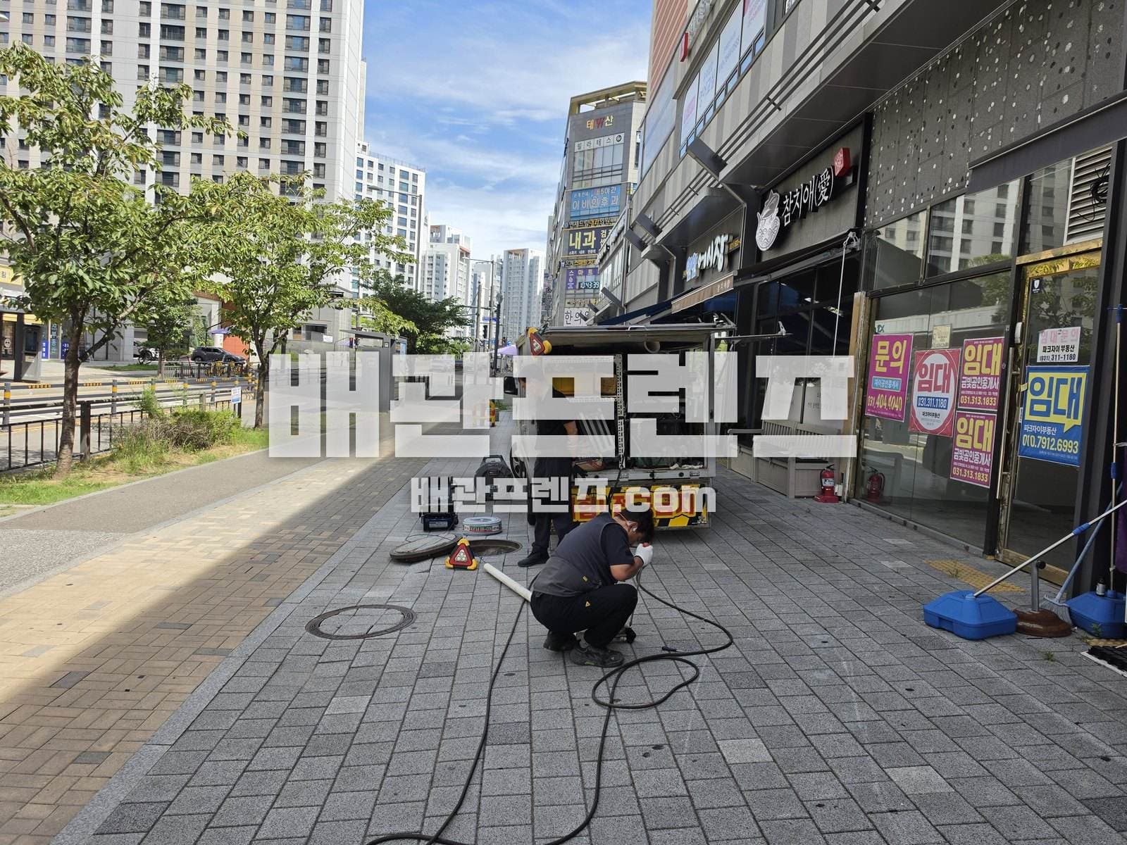 상가 앞 보도 맨홀을 열고 장비 호스를 연결하는 작업자